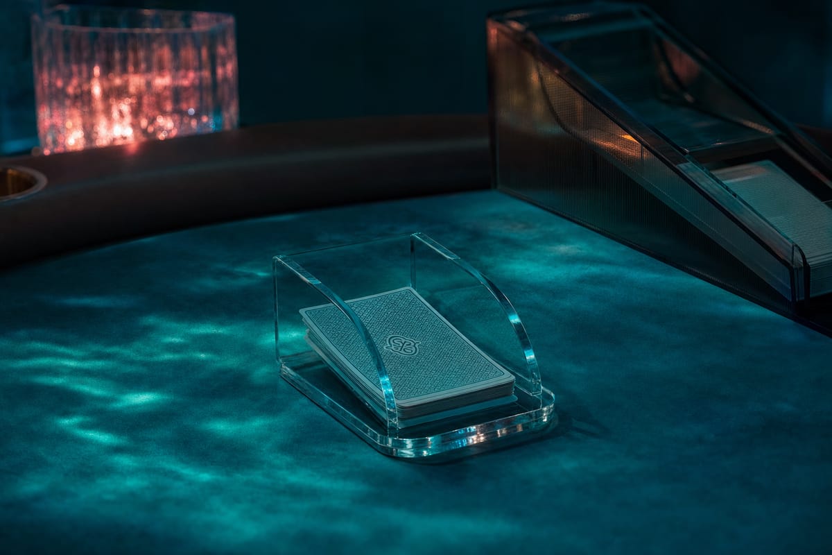 Clear acrylic discard holder with a small stack of used cards, a dealing shoe blurred behind it on teal felt (AI-generated image)