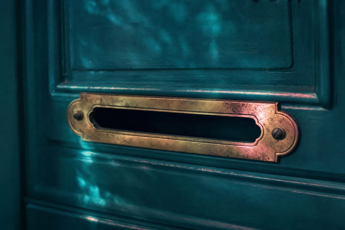 Worn brass letter slot with rounded ends set into a dark teal wooden door, catching warm pink light (AI-generated image)