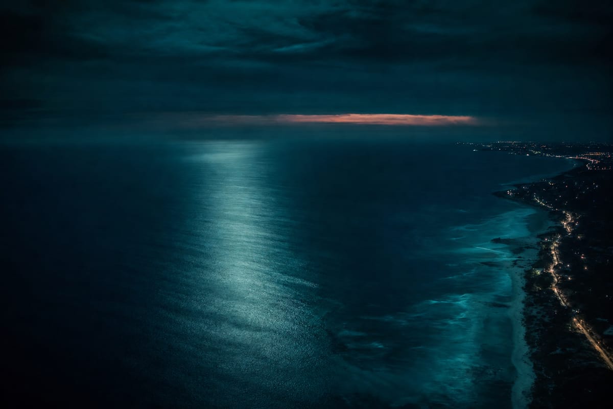 Night view over a dark ocean towards a coastline lit by a thin line of town lights, with a band of orange on the horizon (AI-generated image)