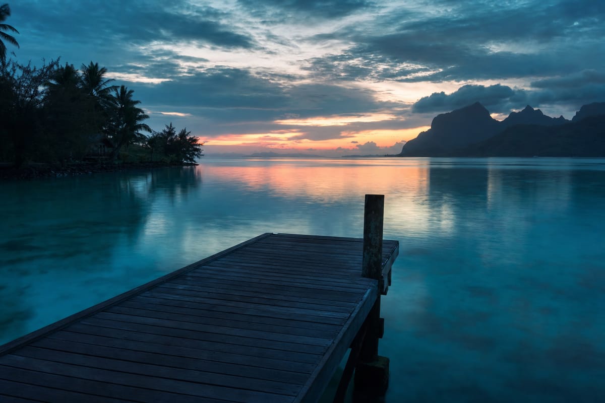 Wooden jetty stretching over a calm lagoon at dusk, with palm trees on the left and mountains on the horizon (AI-generated image)