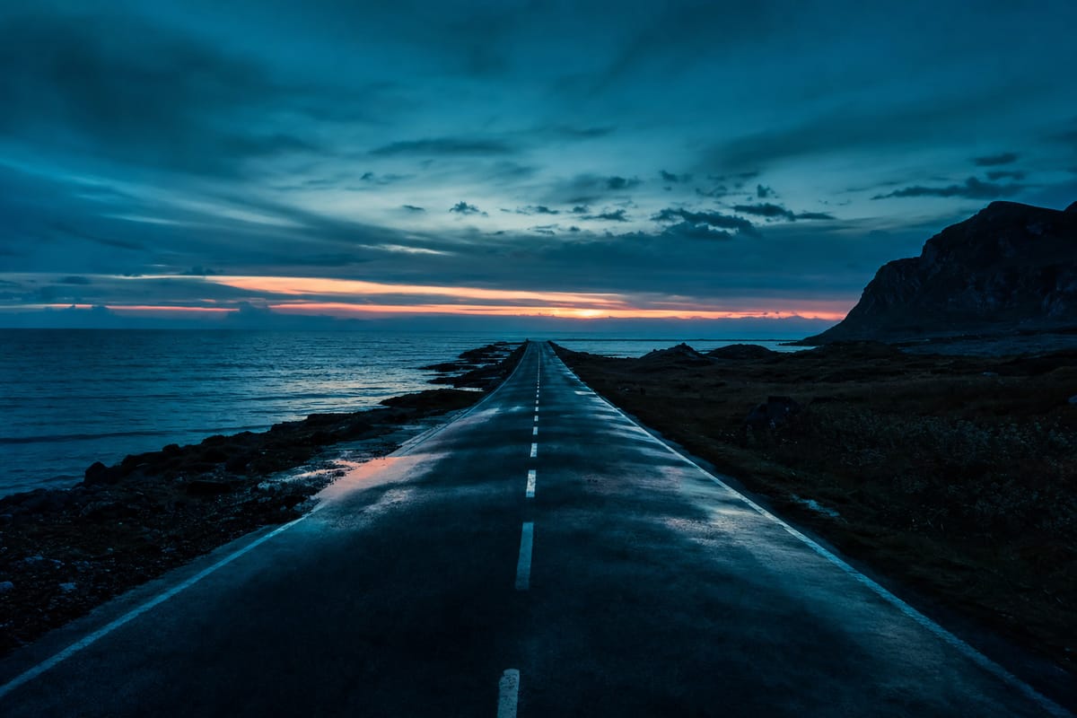 Straight empty coastal road running towards a cliff and the sea under a dusk sky with a thin band of orange light (AI-generated image)