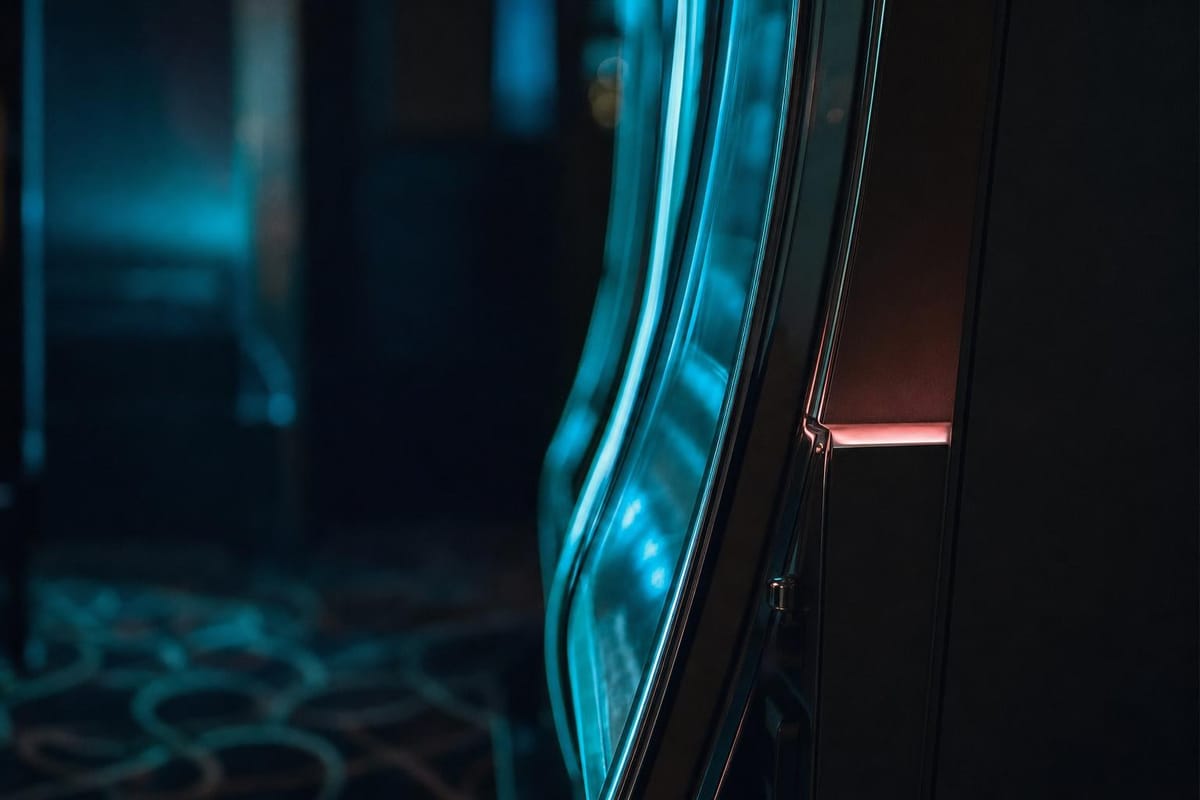Curved chrome and glass side of an upright slot machine cabinet in a dark room, catching teal light and a small coral glow (AI-generated image)
